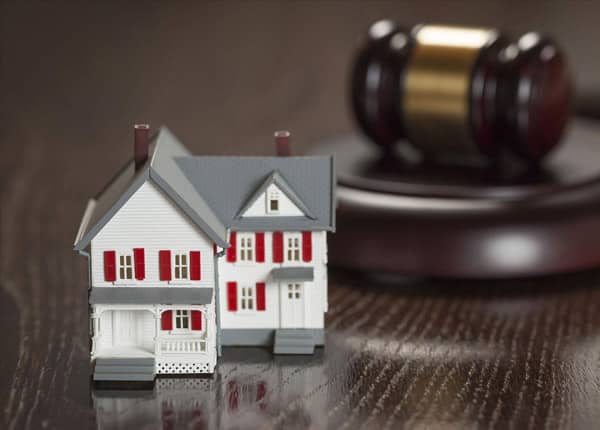An image of a white toy house next to a judge's hammer on a dark brown desk